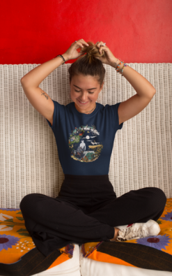 Woman sitting on a couch, tying her hair while wearing a navy blue Studio Ghibli T-shirt with a crescent moon design.