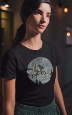 Woman in a dimly lit room, wearing a black T-shirt featuring a Star Wars crossover design with C-3PO and R2-D2 on a bicycle passing the Death Star.