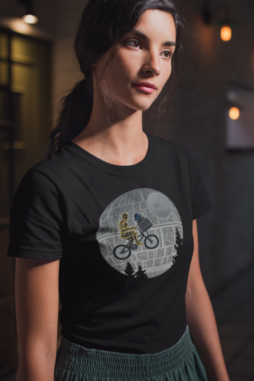 Woman in a dimly lit room, wearing a black T-shirt featuring a Star Wars crossover design with C-3PO and R2-D2 on a bicycle passing the Death Star.