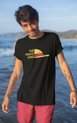 Young man smiling while wearing a black 'Pirate King' T-shirt, standing on a sandy beach with boats in the background.