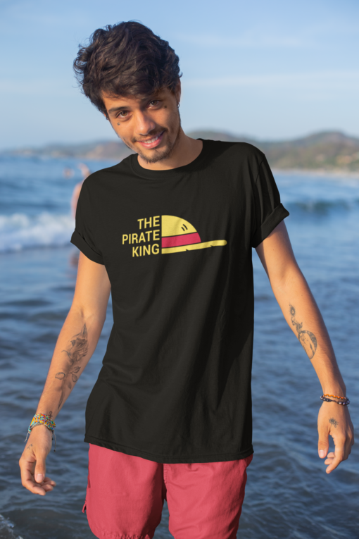 Young man smiling while wearing a black 'Pirate King' T-shirt, standing on a sandy beach with boats in the background.