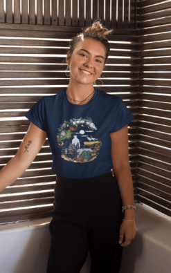 A young woman with a bun and a small tattoo on her arm smiles as she leans against wooden blinds. She is wearing a navy blue T-shirt with a crescent moon-shaped design featuring various animated characters in a scenic blend of nature and fantasy elements.