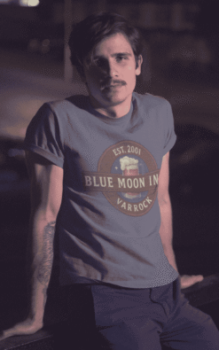 Man modeling Blue Moon Inn Varrock retro t-shirt at night with soft lighting.