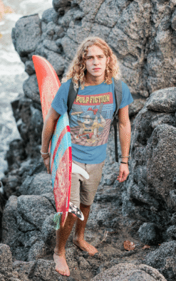Young man wearing Bulma Pulp Fiction mashup t-shirt carrying a surfboard on rocky coastline