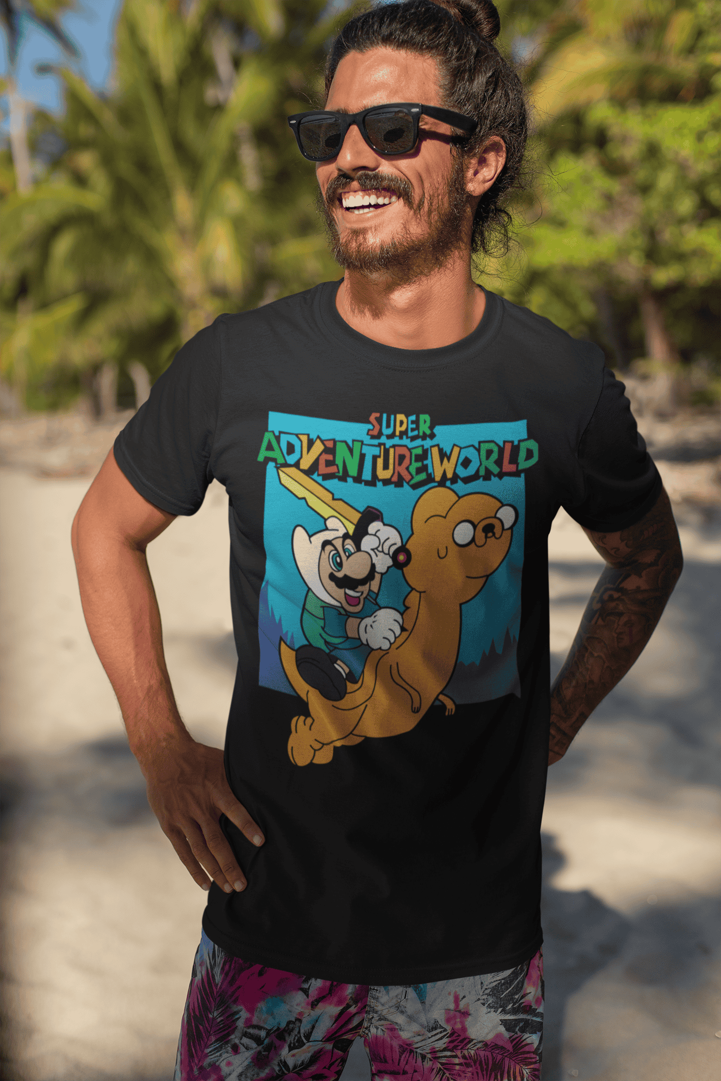 Smiling male model wearing Super Adventureworld black tee with Mario and Jake, sunglasses on, standing on a sunny beach.