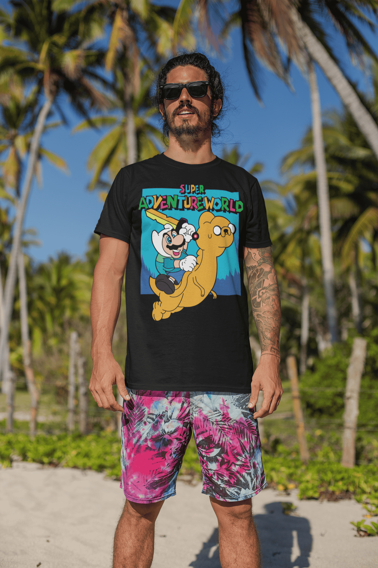 Smiling male model wearing Super Adventureworld black tee with Mario and Jake, sunglasses on, standing on a sunny beach.