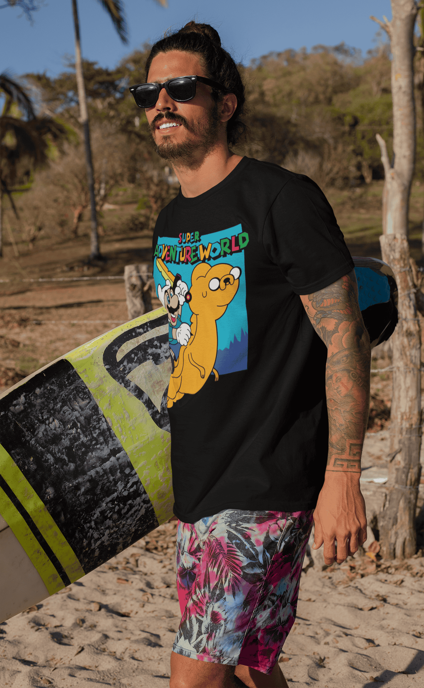 Tattooed male surfer wearing Super Adventureworld black tee featuring Mario and Jake at the beach, smiling confidently.